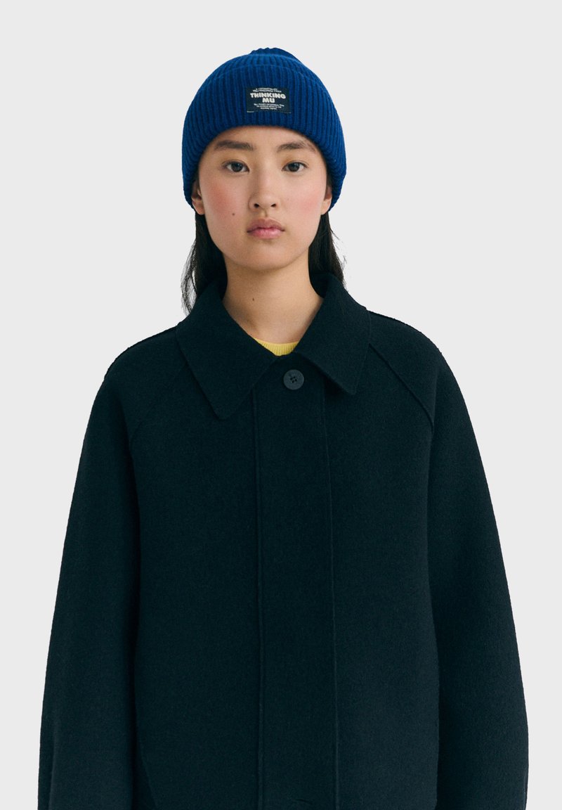 Joven con un gorro de punto azul con un parche y un abrigo negro abotonado sobre una camiseta amarilla, mirando hacia adelante con una expresión neutral.