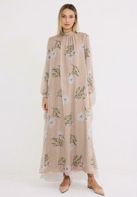 Γυναίκα που φοράει μακρύ μπεζ φόρεμα με λευκά λουλουδένια και πράσινα φύλλα σχέδια, μακριά μανίκια, και μπεζ μυτερά φλατ, στέκεται μπροστά σε λευκό φόντο.