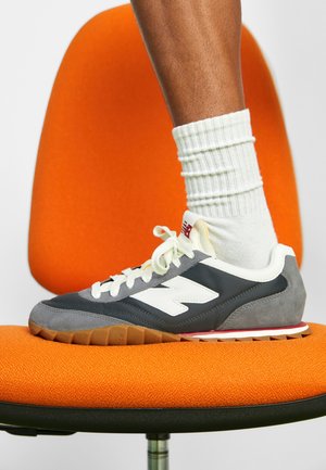 Persona con calcetines blancos y zapatillas New Balance grises de pie sobre una silla de oficina naranja con base negra.