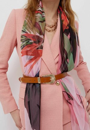 Femme portant un blazer texturé rose, une ceinture en cuir marron avec une boucle dorée, une écharpe fleurie colorée, un collier en chaîne dorée et des boucles d'oreilles assorties.