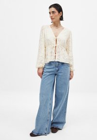 Helles, florales Spitzentop mit langen Ärmeln, gebundenem Vorderteil und V-Ausschnitt. Kombiniert mit weit geschnittenen blauen Jeans und braunen Schuhen.