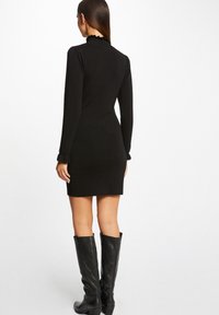 Vestido negro de manga larga con un alto cuello acampanado y silueta ajustada, combinado con botas de cuero negro hasta la rodilla.