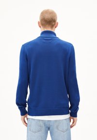 Pull bleu en tricot avec un col montant, à manches longues, bordures et ourlet côtelés. Texture douce, design cintré. Porté par-dessus une chemise blanche et un jean.