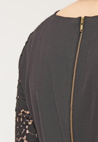 Vestido negro con cierre de cremallera trasero y cremallera dorada. La tela es suave y presenta detalles de encaje en las mangas, mostrando patrones intrincados.