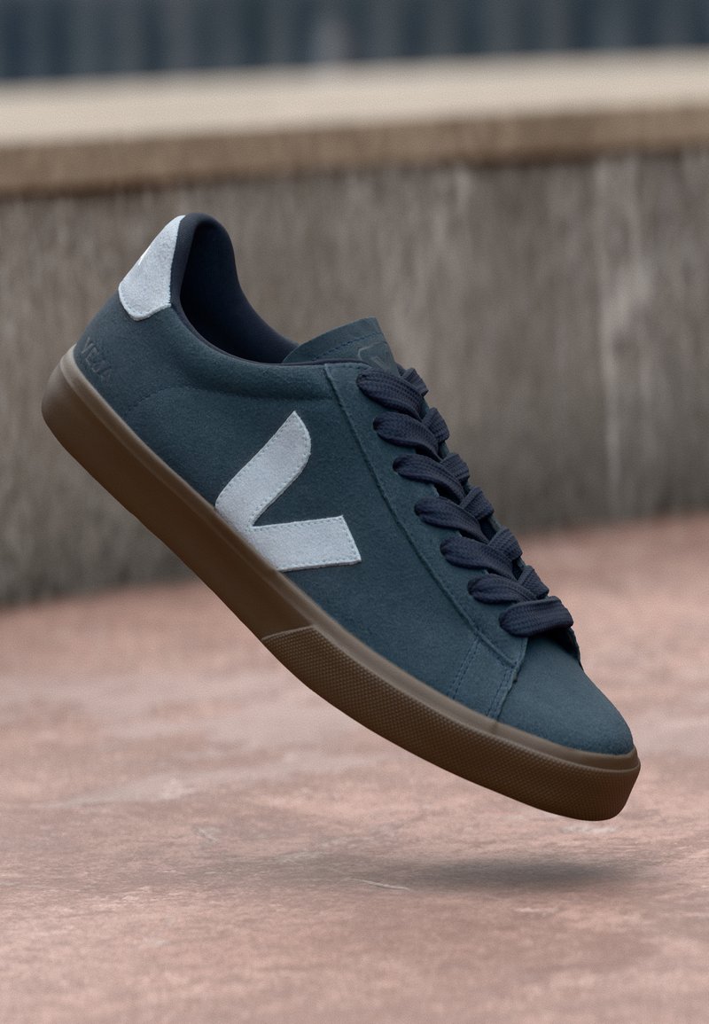 Mėlyni nubuko sportbačiai su balta logo, apvaliu priekiu ir tamsiomis raištelėmis. Gumos padas su tekstūruota danga. Minimalistinis dizainas su kontrastingais akcentais.