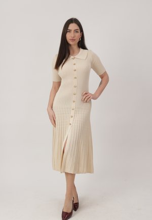 Vrouw met lang donker haar die een crèmekleurige geribbelde midi-jurk draagt met korte mouwen, gouden knopen, kraag en split, gecombineerd met donkerbruine hakken.