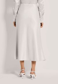 Woman wearing a long, light-colored satin skirt and matching long-sleeve shirt, paired with white high-heeled sandals, shown from the back.