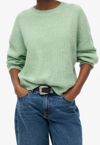 Ljusgrön stickad tröja med avslappnad passform, rund halsringning och uppvikta ärmar, kombinerad med blå högmidjade jeans och ett svart bälte.