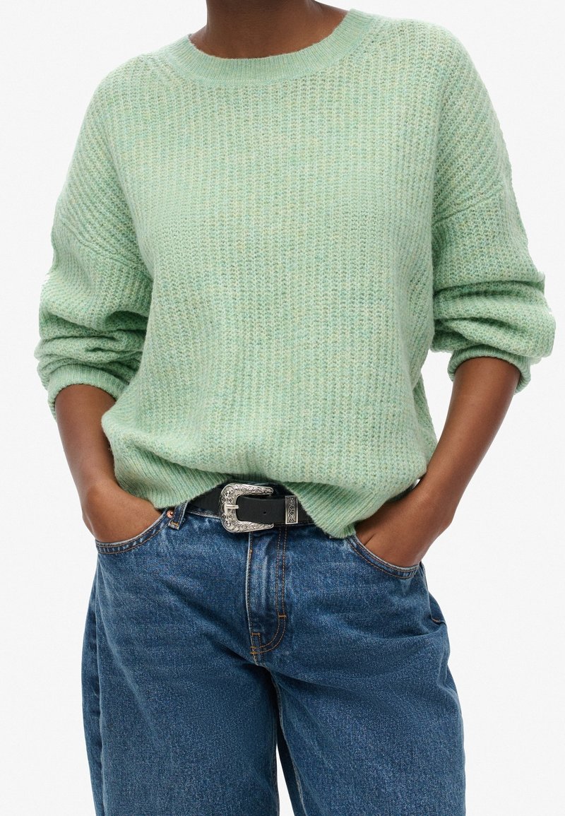 Ljusgrön stickad tröja med avslappnad passform, rund halsringning och uppvikta ärmar, kombinerad med blå högmidjade jeans och ett svart bälte.