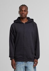 Sudadera con cremallera negra hecha de tejido suave, que cuenta con una gran capucha, bolsillos frontales y puños acanalados. Llevada con jeans desgastados de color azul claro.