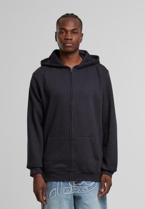 Schwarze Zip-Hoodie aus weichem Material, mit großer Kapuze, vorderen Taschen und gerippten Bündchen. Getragen mit hellblauen, abgetragenen Jeans.