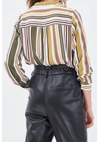 Blouse rayée dans des tons verts, blancs et jaunes ; présente des manches longues, des poignets boutonnés et une coupe décontractée, associée à un pantalon en cuir synthétique noir.