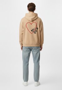 Felpa con cappuccio beige in fleece, decorata con un grande cuore rosso e grafiche di spille di sicurezza nere sul retro. La parte anteriore presenta un cappuccio classico e polsini a coste.