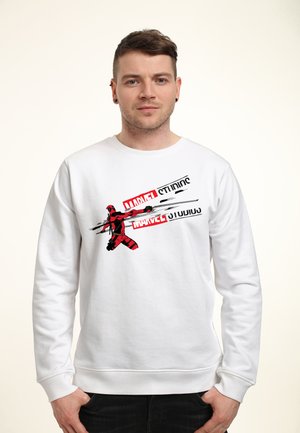 Młody mężczyzna w białej bluzie z czerwono-czarną grafiką Deadpoola i logo Marvel Studios na gładkim jasnym tle.