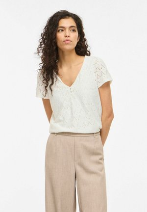 Kobieta o kręconych włosach, ubrana w białą bluzkę z krótkim rękawem z ażurowego materiału i beżowe spodnie z wysokim stanem, stojąca z rękami za plecami.