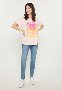Rosa bomulls t-shirt med en grafisk design som föreställer en sol i gult och orange, kombinerad med ljusblåjeans och vita sneakers.