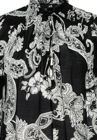 Schwarze Bluse aus Stoff mit einem weißen Blumen- und Paisleymuster, die mit einem gerafften Ausschnitt und einer Schleifendetail versehen ist. Glatte Textur mit fließendem Design.