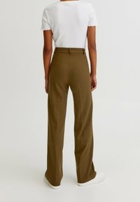 Persoon in een hoge taille olijfgroene wijde broek, wit T-shirt met korte mouwen en witte sneakers, staand met de rug naar de camera op een effen achtergrond.