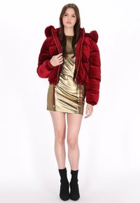 Chaqueta de plumón de terciopelo rojo con capucha, puños acanalados y detalles de cremallera, combinada con un vestido ajustado de metal dorado y botas negras de tobillo.