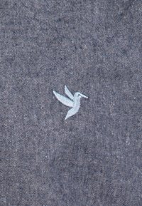Colibrí azul bordado en una tela gris texturizada, con un fino bordado y un patrón sutil, posicionado de manera prominente en el material.