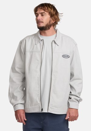 Light gray denim jacket with a zip front, pointed collar, and elastic cuffs. Features a logo patch on the left chest. Soft texture.
