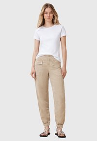 Bílý bavlněný tričko v kombinaci s béžovými cargo kalhotami s předními kapsami, volným střihem a elastickými manžetami, doplněné stříbrnými sandály.