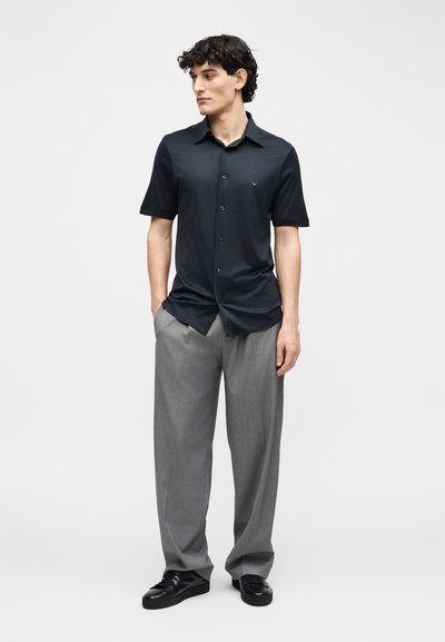 Νεαρός άνδρας όρθιος, φοράει μαύρο πουκάμισο με κοντά μανίκια και κουμπιά, άνετο γκρι παντελόνι και μαύρα παπούτσια, κοιτάζει προς τα πλάγια.
