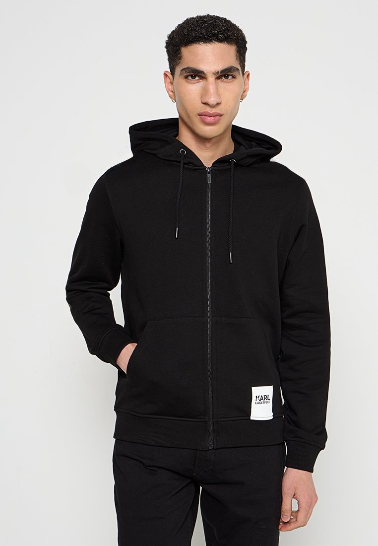 Sudadera negra con cremallera hecha de un tejido suave, que presenta un bolsillo frontal, capucha con cordones, y una etiqueta cosida en la parte inferior derecha.