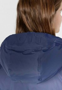 Giacca a vento blu navy con una texture liscia e lucida; logo "JOTT" in rilievo sul retro; presenta un design pratico e funzionale.