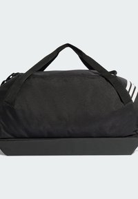 Bolsa de lona negra hecha de material duradero, con dos asas superiores, forma rectangular y tres rayas blancas en un lado.