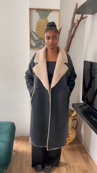 Abrigo largo negro con un cuello de piel sintética beige, diseño de doble botonadura y costuras visibles. Lleva puesto sobre una blusa y pantalones negros.