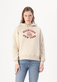 Felpa crema con stampa grafica frontale che recita "HILFIGER est 1985 NEW YORK." Caratterizzata da cappuccio con cordino e polsini a costine, abbinata a jeans blu.