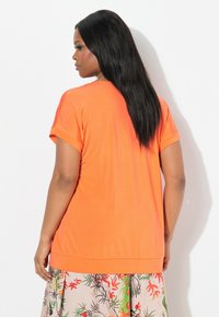 T-shirt arancione a maniche corte con una vestibilità rilassata e risvolti, abbinato a una gonna floreale a fantasia con tonalità di beige, verde e rosso.