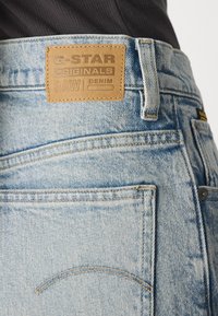 Svetlomodré džíny z denimu s hnedou koženou nášivkou s embosovaným textom, klasický dizajn s piatimi vreckami a jemné detaily vyblednutia.