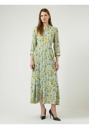 Kvinne iført en lys blå, lang blomsterkjole med gule blomster, knapper foran, trekvart ermer og svarte høyhælte sko.