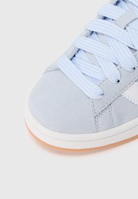 Světle modrá semišová teniska s texturovaným povrchem, plochými tkaničkami a bílou gumovou podrážkou s jemným oranžovým akcentem na spodní straně.