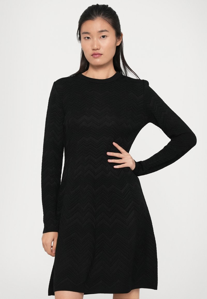 Robe longue noire à manches longues avec un motif zigzag texturé, dotée d'un col rond et d'une silhouette évasée. Tissu lisse, longueur au-dessus du genou.