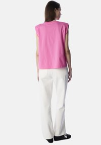 Roze mouwloze top met schoudervulling, gecombineerd met witte brede broek en zwarte schoenen, gezien vanaf de achterkant.