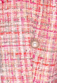 Vaaleanpunainen ja korallin sävyinen teksturoidusta kankaasta valmistettu, jossa on kudottu kuvio ja hopeanvärinen nappi, jonka keskellä on helmi, mikä toimii merkittävänä yksityiskohtana.