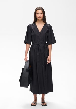 Femme debout portant une robe portefeuille noire avec des manches aux coudes, tenant un grand sac à main noir et portant des sandales noires.