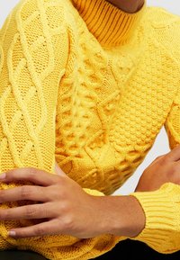 Pull à col roulé jaune tricoté avec motifs texturés, torsades et poignets côtelés. Vue rapprochée mettant en valeur les détails du matériau et du design.