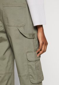 Olive green cargo pants with multiple pockets, loop details, visible stitching, and a smooth, durable fabric; hand resting on the side pocket.