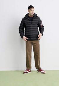 Giacca a vento nera con cappuccio, design aderente, indossata sopra una maglietta nera, abbinata a pantaloni marroni e sneakers bordeaux. Sfondo semplice.