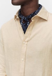 Beige liinapaita, jossa on napitettava edusta ja tummansininen bandana kaulalla, joka esittää valkoisia kukkakuvioita ja aivan kuten solmitun muodon.