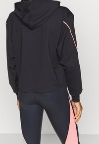 Felpa nera corta con cappuccio, con strisce rosa a contrasto sulle maniche. Indossata con leggings lucidi e aderenti con un dettaglio a striscia in contrasto.