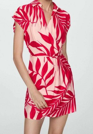 Mujer con un vestido de manga corta y cuello, con un estampado de hojas rojas sobre un fondo rosa claro, de pie frente a un fondo liso.