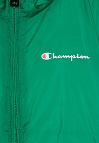 Žalias nailoninis švarkas su pilnu užtrauktuku, lygia tekstūra ir baltu "Champion" logotipu priekyje. Lengvas ir oro pralaidus dizainas.