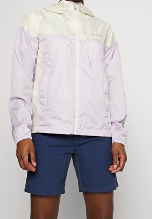 Person iført en lyse lilla og cremefarvet Columbia lynlåsvindjakke og marineblå shorts, stående mod en ensfarvet baggrund.