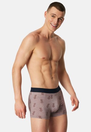 Giovane uomo muscoloso che sorride, indossa boxer viola con stampa di ananas scuro e una fascia in vita nera, in piedi davanti a uno sfondo semplice.