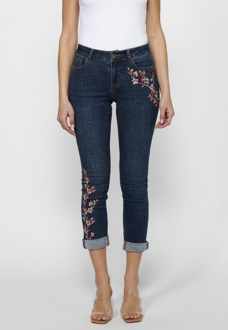 Jeans de mezclilla en azul oscuro con bordado floral en el lateral. Presentan un corte ajustado, puños enrollados y un cierre de botón tradicional.